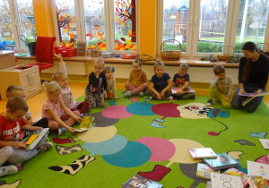Grupa dzieci siedzi na dywanie wokół rozłożonych książek, niektóre z dzieci trzymają w ręku książkę, w której oglądają ilustrację.